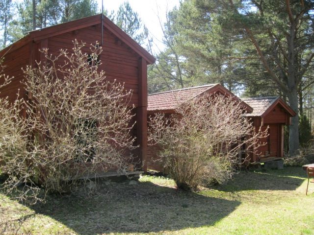 Röd timmerbyggnad med flera bodar i rad, omgiven av buskar och träd.