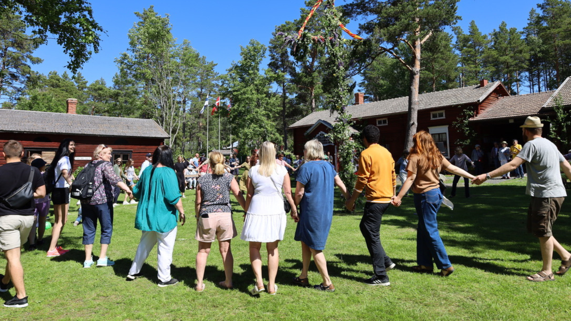 Besökare dansar runt midsommarstång på Kilens hembygdsgård i soligt väder